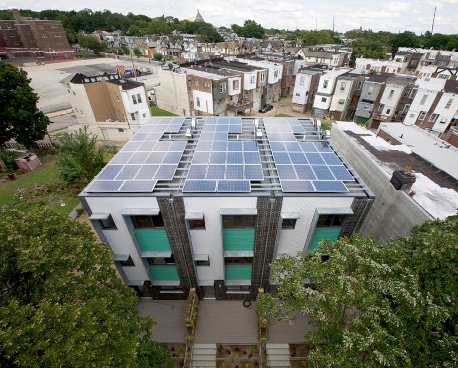 Modern, sustainable multifamily housing; ariel shot showing solar panels on roof, trees and rowhomes surrounding