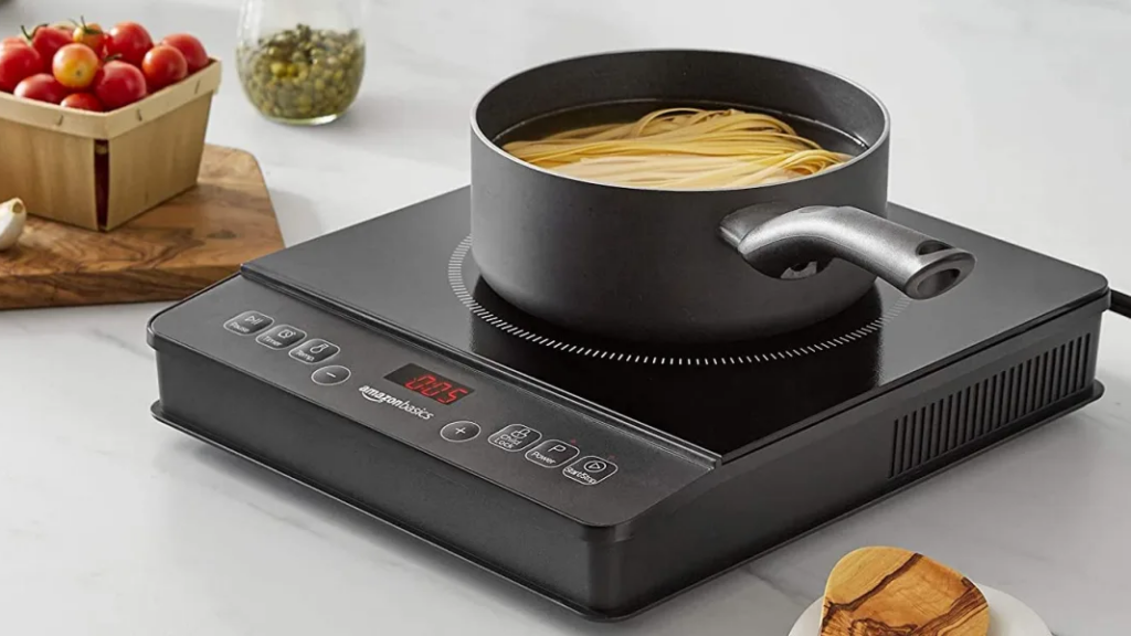 Black pot containing water and noodles sits atop induction burner on white backgroud and some food items surrounding - photo
