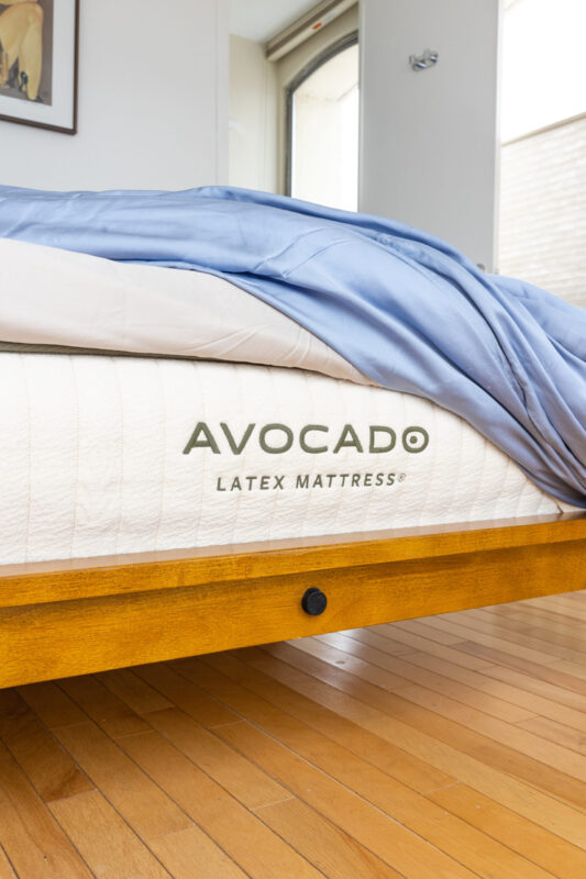 Close shot of foot of mattress showing Avocado logo and bedding; sits on wood bedframe and wood flooring in room with white wall and large shaded windows - photo