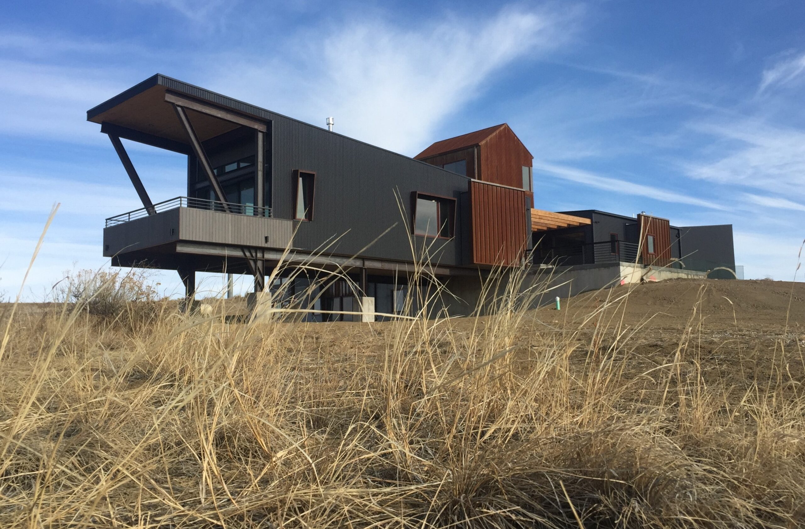 exterior view of modern house; dark siding and striking, cantilevered geometries; dense dry grass in foreground; blue sky in background - photo