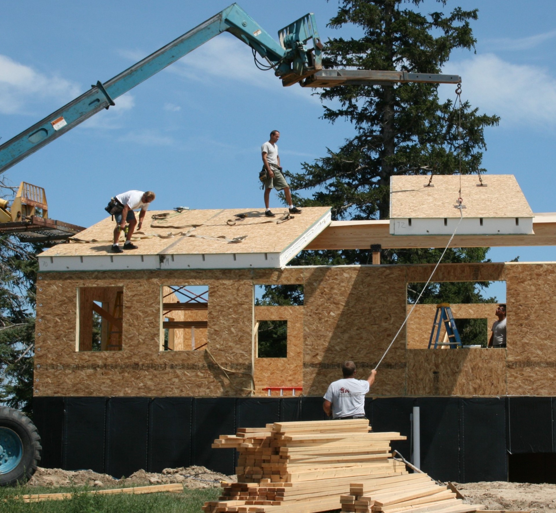 exterior view of home under construction using SIP panels; workers on the ground and on the roof; loader lowers large insulated roof panel into position - photo