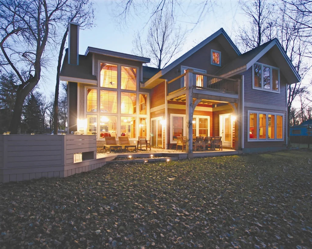 exterior of timber-frame home built with structural insulated panels; warm interior lighting sines from huge windows; setting shows bare trees and fallen leaves on the lawn - photo