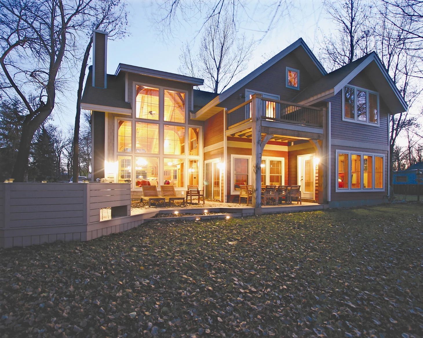exterior of timber-frame home built with structural insulate panels; warminterior lighting sines from huge windows; setting shows bare trees and fallen leaves on the lawn - photo