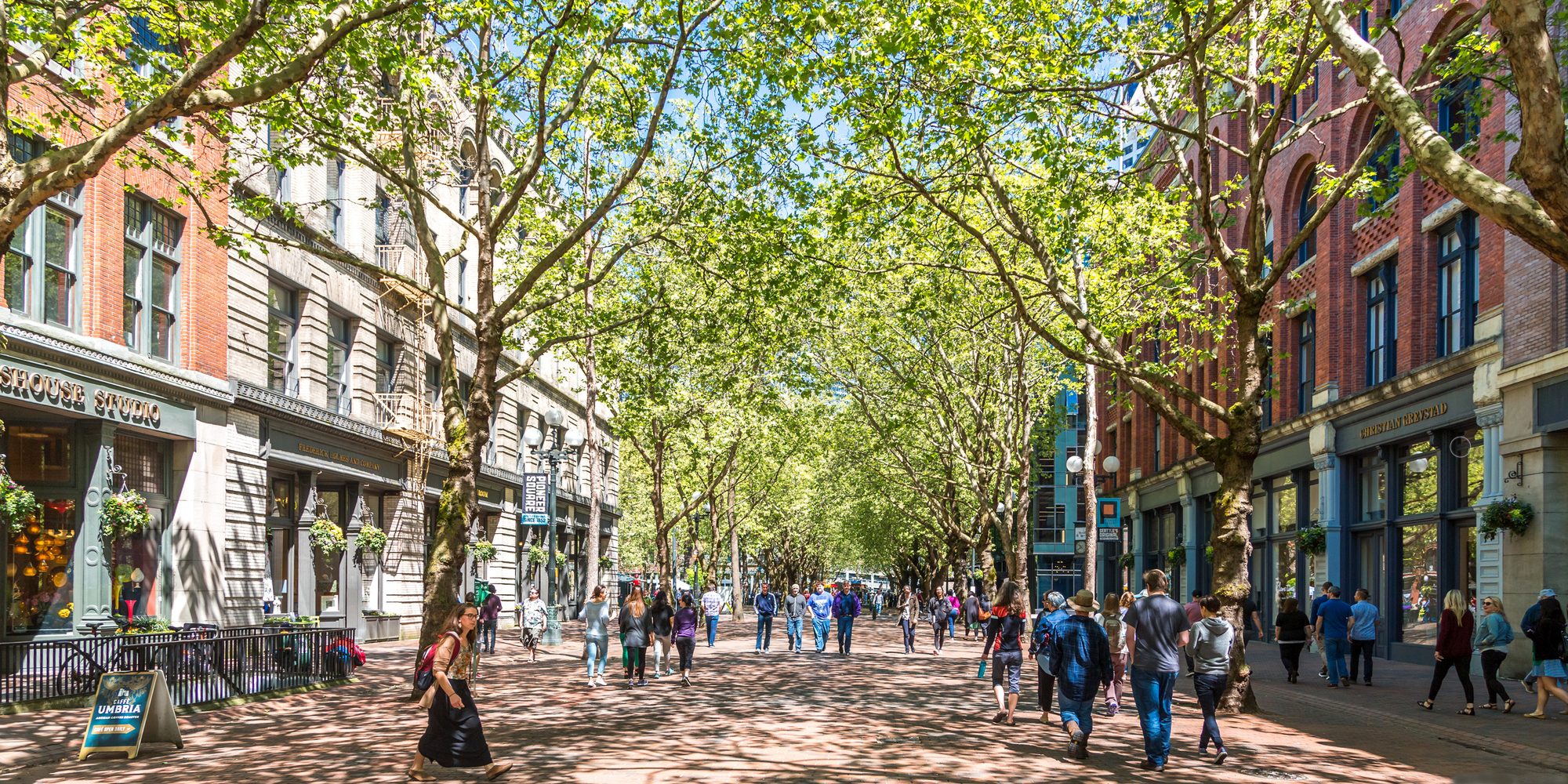 walkable community in Seattle shows view down street blocked off from traffic, with pedestrians walking on the street and sidewalks; green leafy trees arch overhead; mixed-use development lines the street - photo